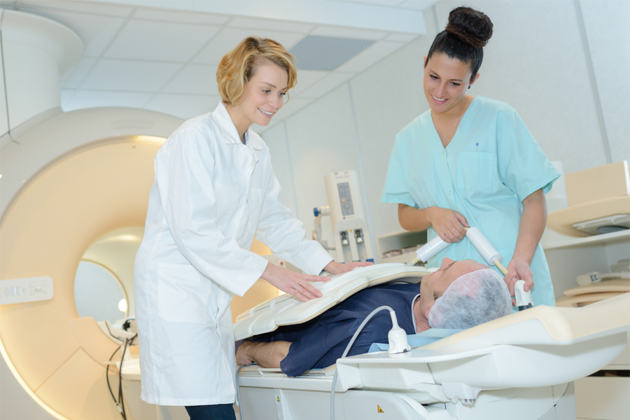 Imagem: dupla formada por uma enfermeira e uma técnica de radiologia especialista em medicina nuclear acalmam e preparam paciente para realização de exame radiológico.