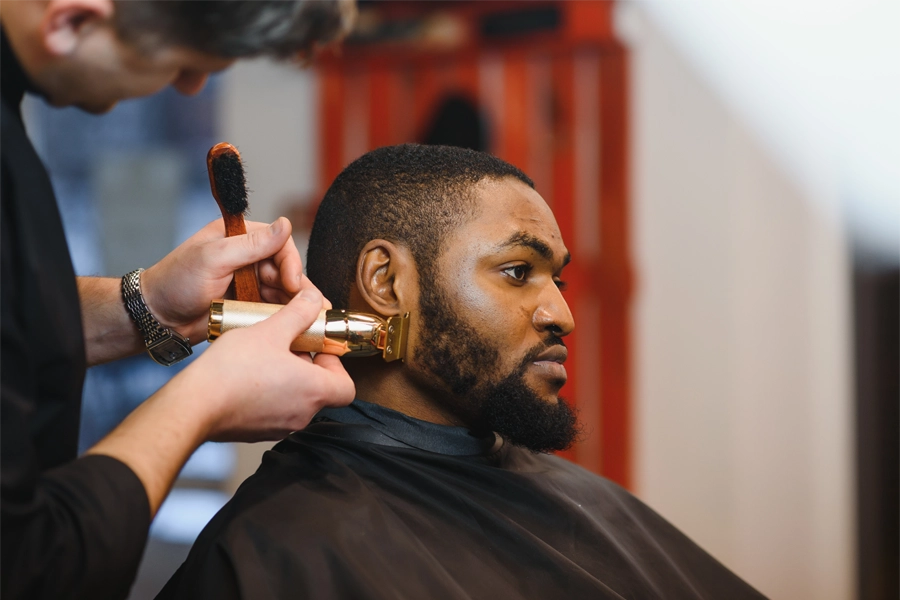 Barbeiro finalizando a barba de um cliente com a ajuda de uma máquina para barba e um pente.