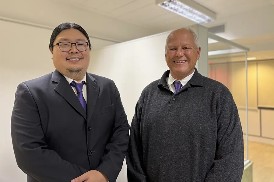 Aldo e Francisco estão sorrindo e utilizando traje formal preto com gravata roxa.