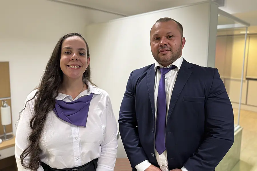 Carolina e Gilmar estão sorrindo, utilizando traje formal com lenço e gravata roxa.
