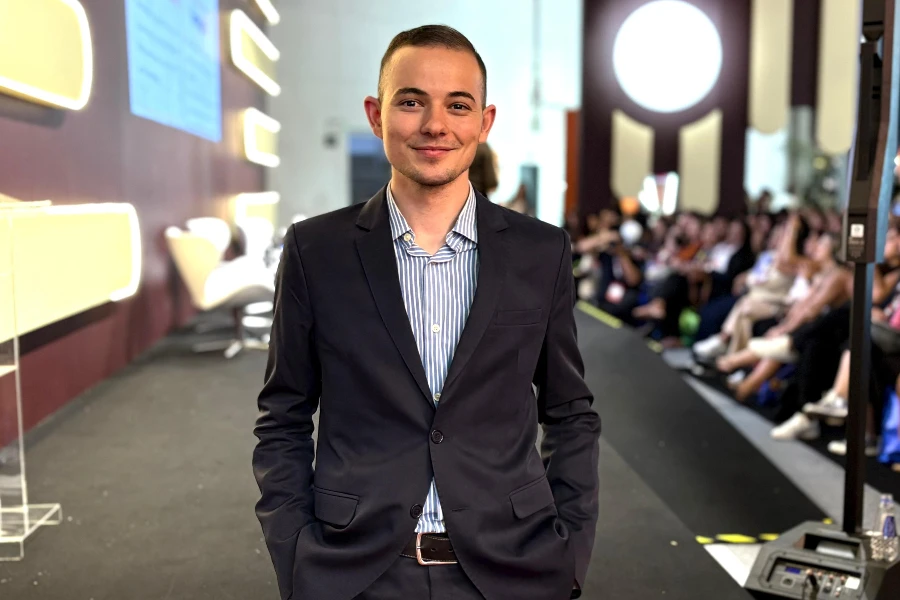 David está sorridente com as mão no bolso, ele usa terno e está no palco de uma palestra