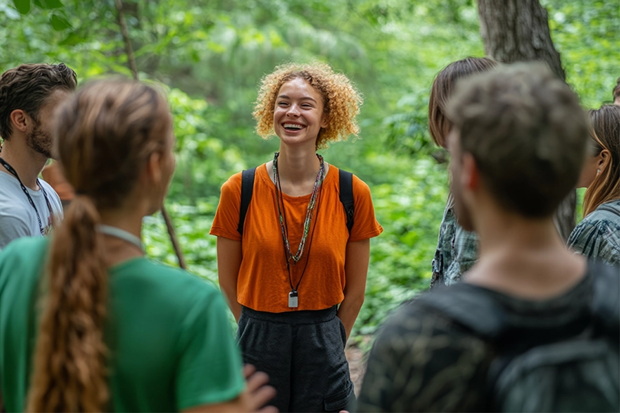 Grupo de seis pessoas em área arborizada. No centro, uma guia de turismo com camiseta laranja e colar com apito, cercada por outras pessoas, algumas de costas para a câmera
