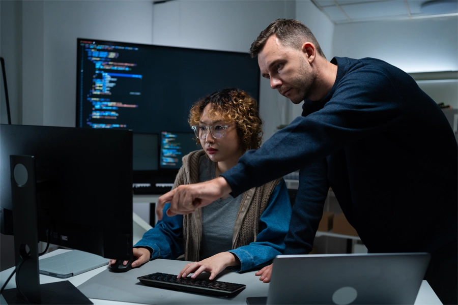 Imagem: dois colegas de trabalho da área de tecnologia, uma mulher jovem e um homem jovem, trabalham juntos em uma tarefa de tecnologia da informação.