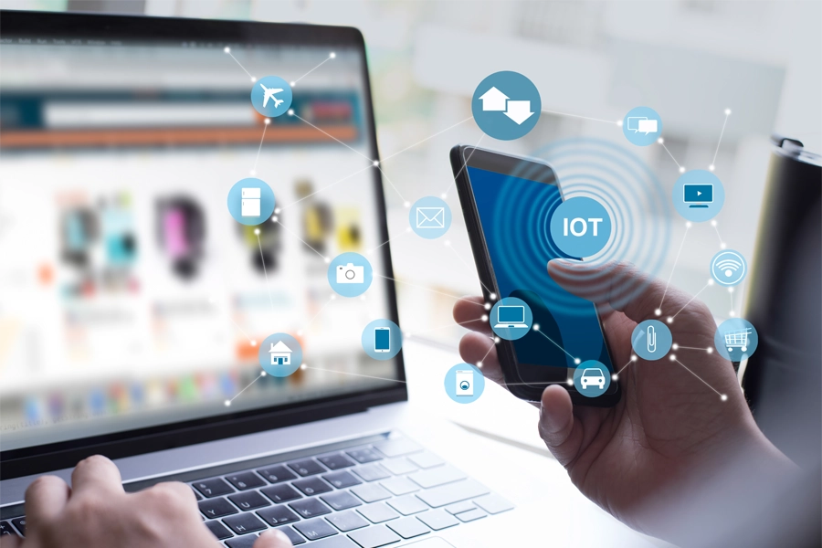 Imagem: a mão de uma pessoa é exibida segurando um celular com a sigla "IoT" conectada a figuras de computador, carro, clips de papel e casa simulando uma vida com dispositivos conectados.
