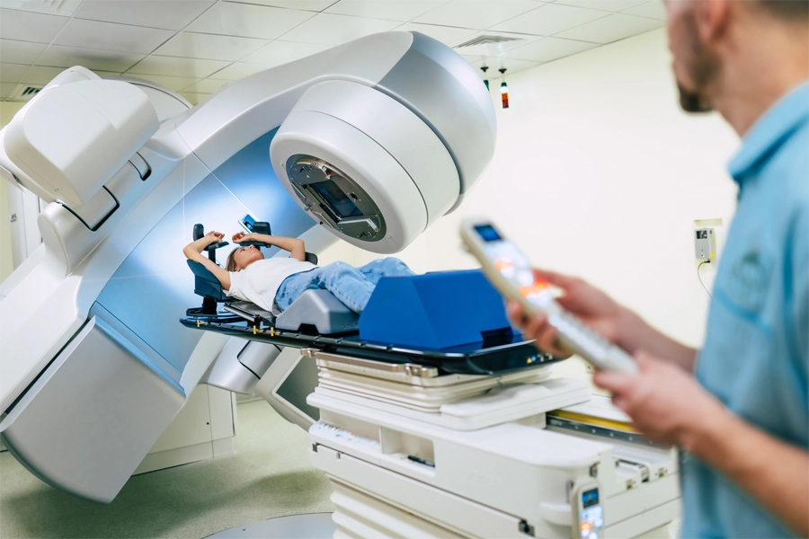Imagem: paciente apoia seus braços em máquina de tomografia enquanto profissional de radiologia ajusta os controles para a realização do exame.