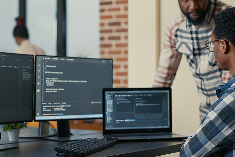 Professor negro em pé, ao lado de aluno negro ensinando códigos de programação.