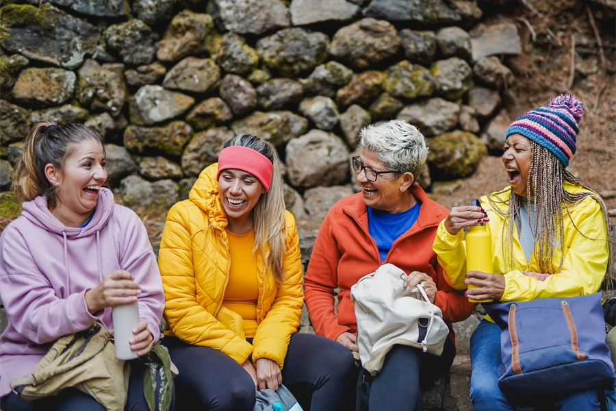 Imagem: grupo de mulheres de diferentes idades sorriem sentadas uma ao lado da outra na pausa de uma trilha. Atrás delas, uma parede de pedras.