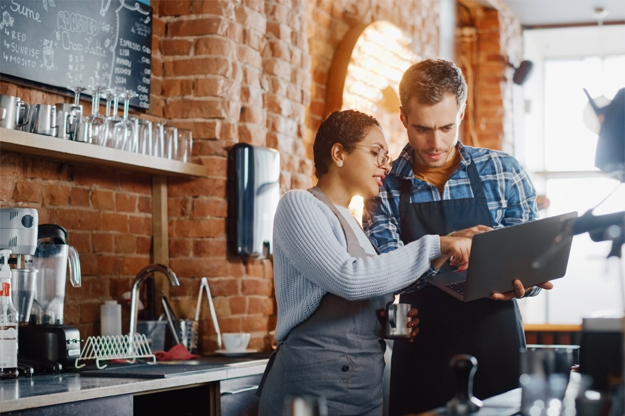 Imagem: dois técnicos em serviços de restaurante e bar conversando no local de trabalho enquanto consultam informações na tela de um notebook.