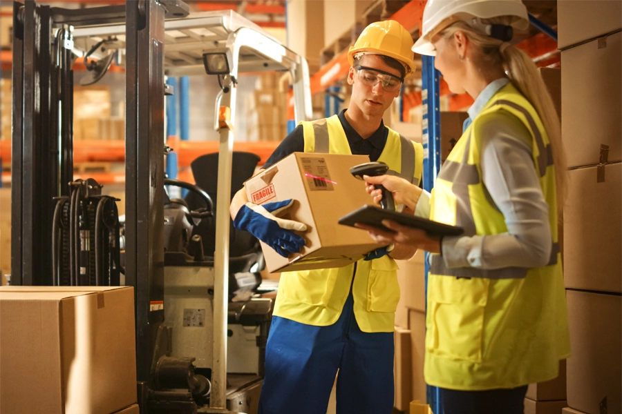 Imagem: dois técnicos em logística vestidos com roupas de segurança fazem o registro de mercadorias em um depósito de produtos.