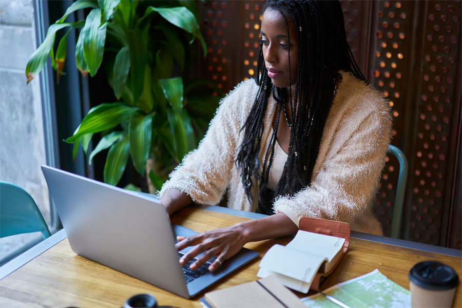 Uma mulher negra, com tranças soltas na altura do ombro e usando um casado peludo de cor clara está trabalhando em um computador apoiado a uma mesa de madeira.