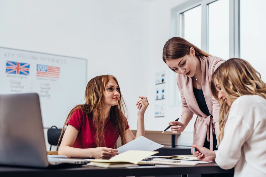 professora auxilia duas alunas que estão sentadas fazendo uma atividade na mesa que contém cadernos e laptop. Ao fundo, há um quadro branco com bandeiras do Reino Unido e dos Estados Unidos e referências de ortografia em inglês.