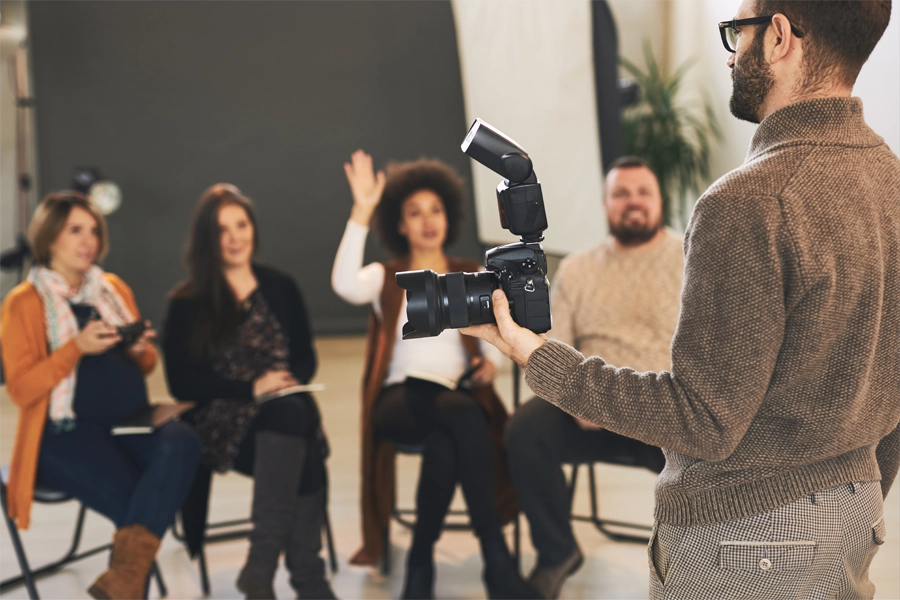 Imagem: estudantes de fotografia sentados e em roda durante aula do curso, o professor está em pé segurando uma câmera e pronto para atender a uma aluna que sinaliza dúvida com o braço levantado.