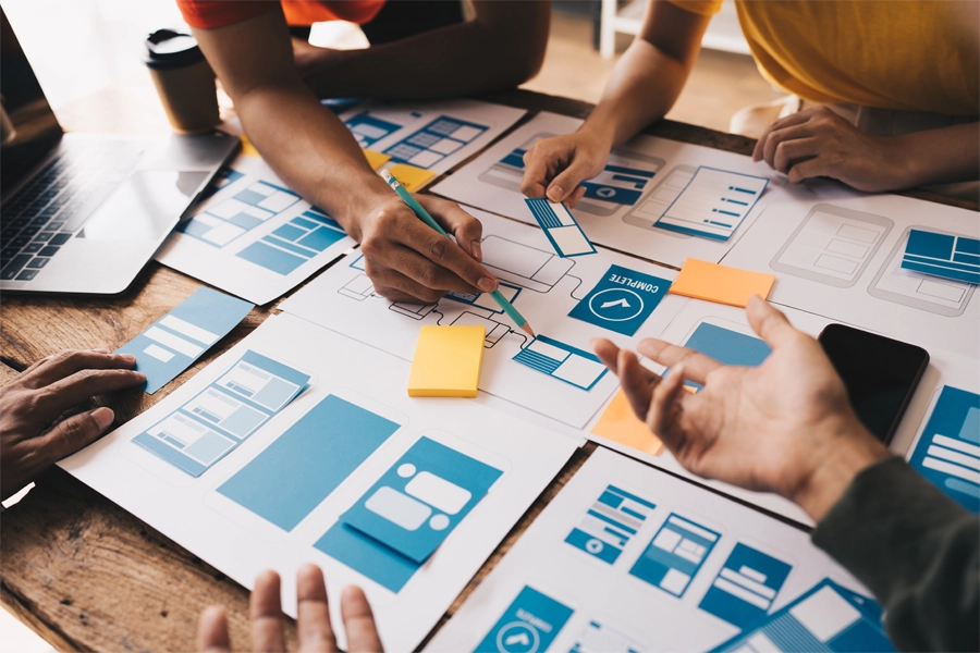 Imagem: uma mesa com folhas de papel com ícones e colunas em diferentes formatos é exibida junto as mãos de pessoas apontando e debatendo sobre as escolhas um projeto UX.