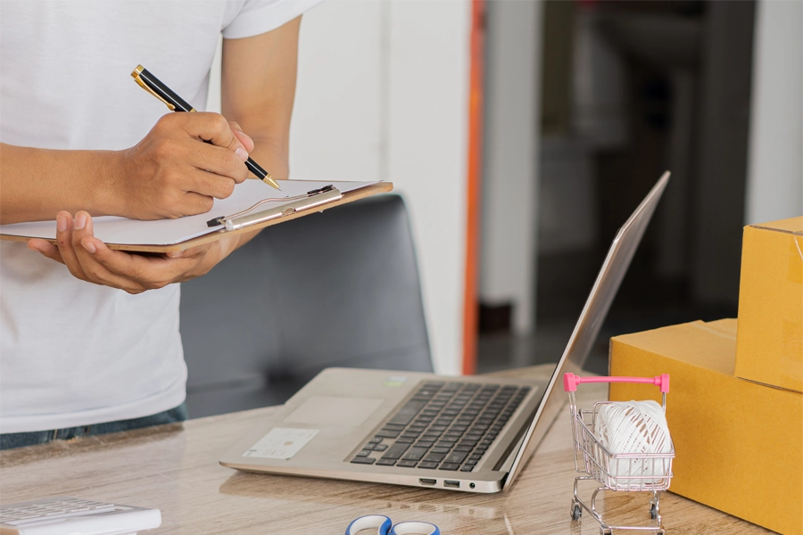 Imagem: um homem utiliza uma prancheta para anotar informações checadas na tela de um notebook apoiado em mesa de escritório.