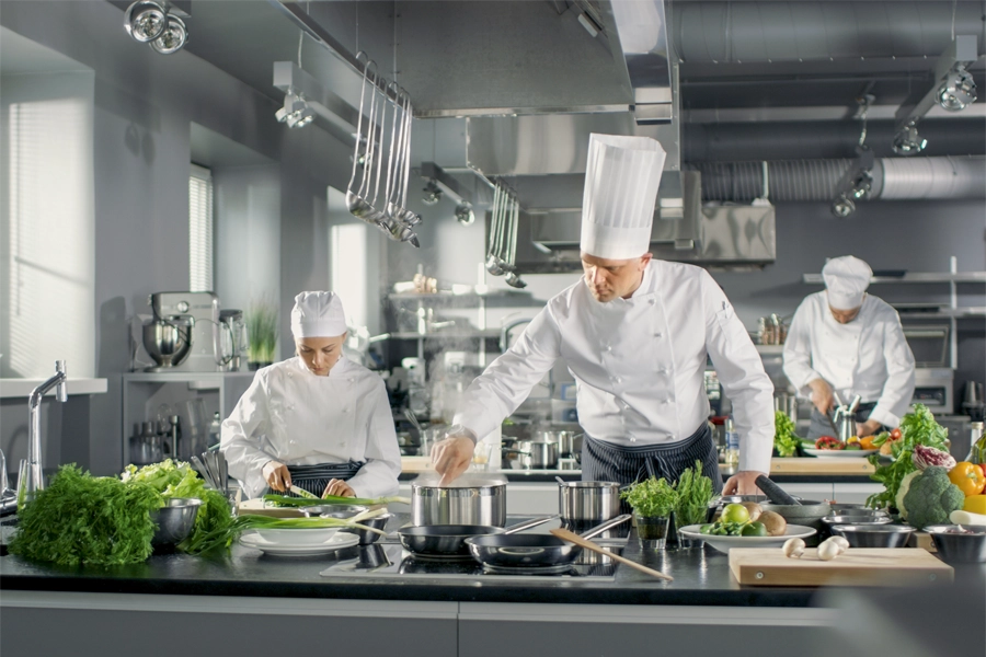 Imagem: três chefs de cozinha dividem bancada de cozinha industrial durante preparos culinários.
