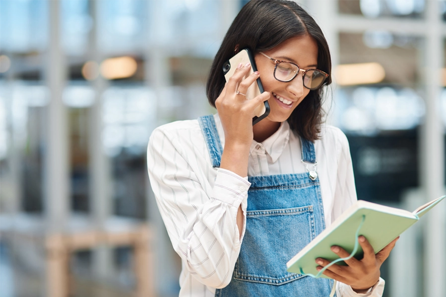 Imagem: uma mulher técnica em eventos olha para as anotações de um caderno enquanto fala ao telefone.