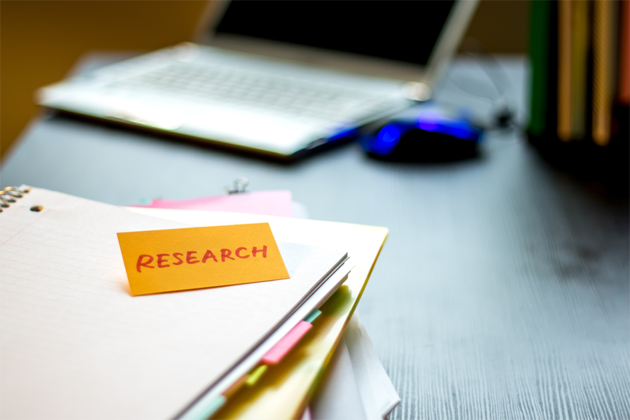 Mesa com caderno e folhas organizadas com marcadores coloridos, sobre as quais há um cartão laranja com a palavra 'RESEARCH' escrita em letras maiúsculas. Ao fundo, um notebook aberto e um mouse azul sobre a mesa.