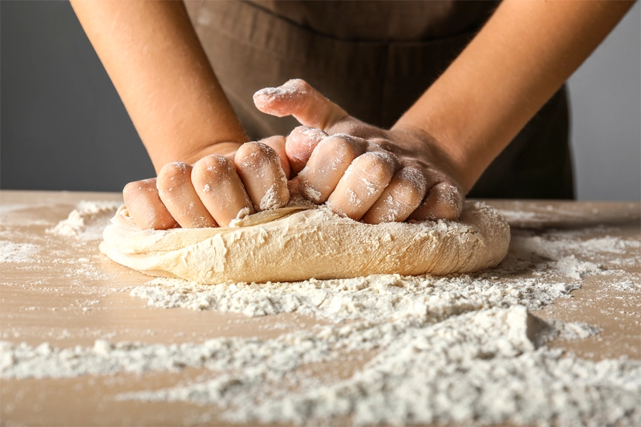 Imagem: close de duas mãos sovando uma massa de pão com farinha de trigo apoiando em uma mesa de madeira.