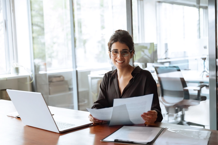 Imagem: uma técnica administrativa está sentada com papéis na mão, olhando para a frente e sorrindo.