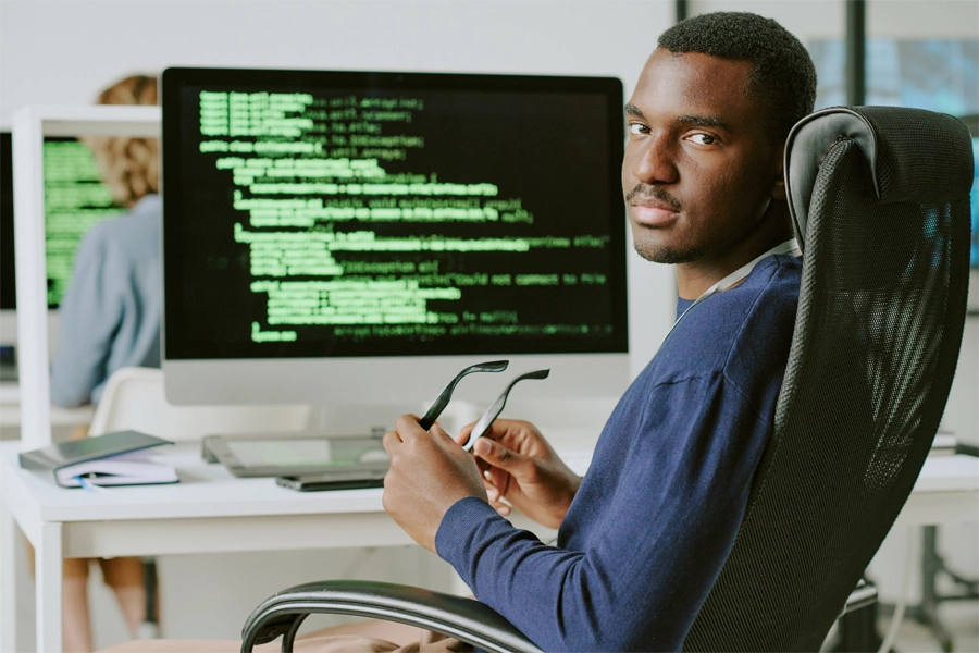 Imagem: um analista em segurança cibernética aparece sentado em sua mesa de trabalho, onde uma tela é exibe códigos de programação enquanto ele se vira e olha para trás segurando seus óculos nas mãos.