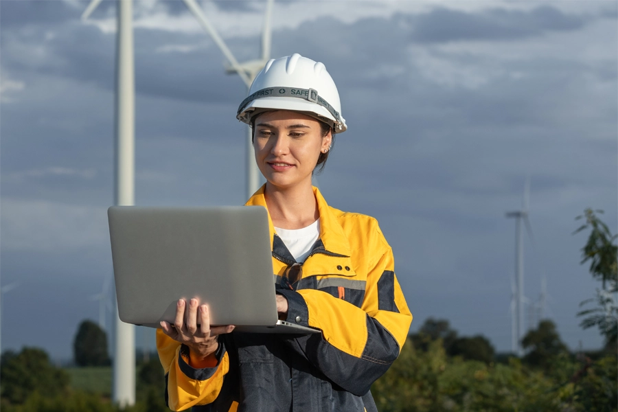 Imagem: em um espaço a céu aberto, uma técnica em meio ambiente uniformizada confere informações na tela de um notebook aberto. Atrás dela é possível ver aerogeradores de energia eólica.