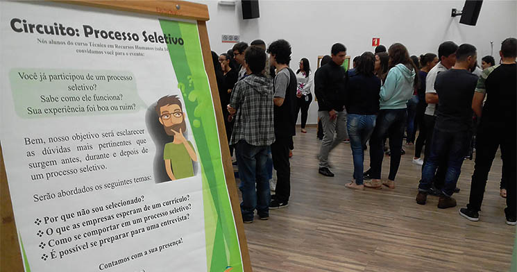 Banner sobre o Circuito Processo Seletivo com público ao fundo na sala Banner sobre o Circuito Processo Seletivo com público ao fundo na sala