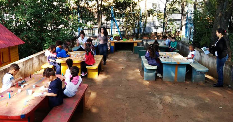 Duas alunas do Técnico em Meio Ambiente interagem com crianças em um espaço aberta da escola municipal com mesas e cadeiras coloridas feitas de concreto durante atividade educativa Duas alunas do Técnico em Meio Ambiente interagem com crianças em um espaço aberta da escola municipal com mesas e cadeiras coloridas feitas de concreto durante atividade educativa