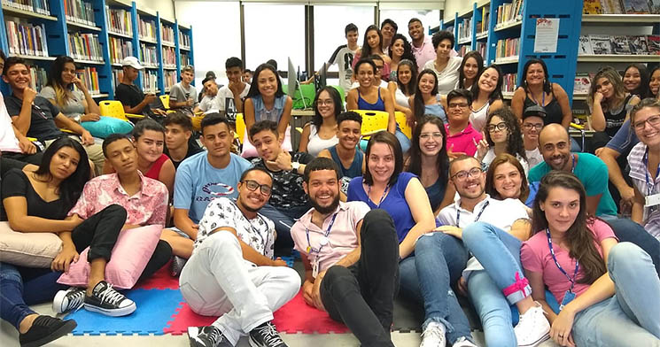 Docente Daniel Camargo sentado no chão da biblioteca ao lado dos jovens aprendizes Docente Daniel Camargo sentado no chão da biblioteca ao lado dos jovens aprendizes
