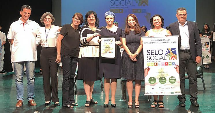 Oito pessoas sorridentes no palco do evento de premiação. Uma delas segura um banner com logo e nome do programa Selo Social e outra segura uma placa do selo Oito pessoas sorridentes no palco do evento de premiação. Uma delas segura um banner com logo e nome do programa Selo Social e outra segura uma placa do selo