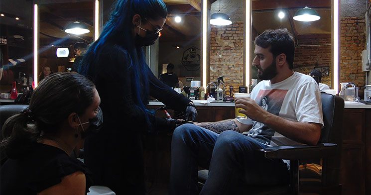 Homem sentado em cadeira de barbearia com cerveja na mão enquanto recebe serviços de manicure e pedicure de duas mulheres Homem sentado em cadeira de barbearia com cerveja na mão enquanto recebe serviços de manicure e pedicure de duas mulheres