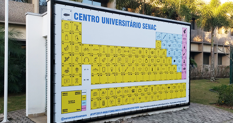 Foto em área externa, em dia de sol, com uma tabela periódica gigante em um painel amarelo Foto em área externa, em dia de sol, com uma tabela periódica gigante em um painel amarelo