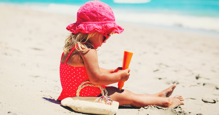Foto de uma menina com maiô vermelho de bolinhas brancas, chapéu rosa e óculos de sol sentada na areia da praia passando filtro solar nas pernas Foto de uma menina com maiô vermelho de bolinhas brancas, chapéu rosa e óculos de sol sentada na areia da praia passando filtro solar nas pernas
